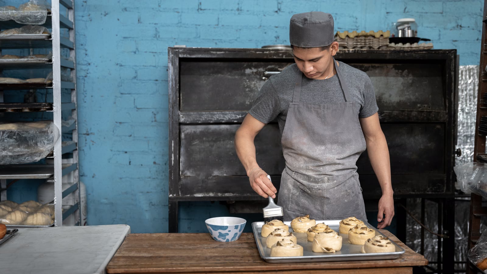 A baker glazing a tray of rolls at a worn wooden table in front of an iron oven.