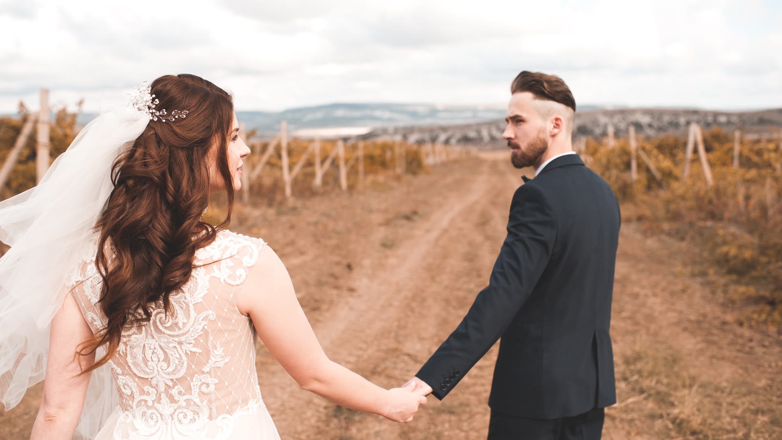 A couple in wedding clothes walking a dirt track between vine rows, hand in hand.