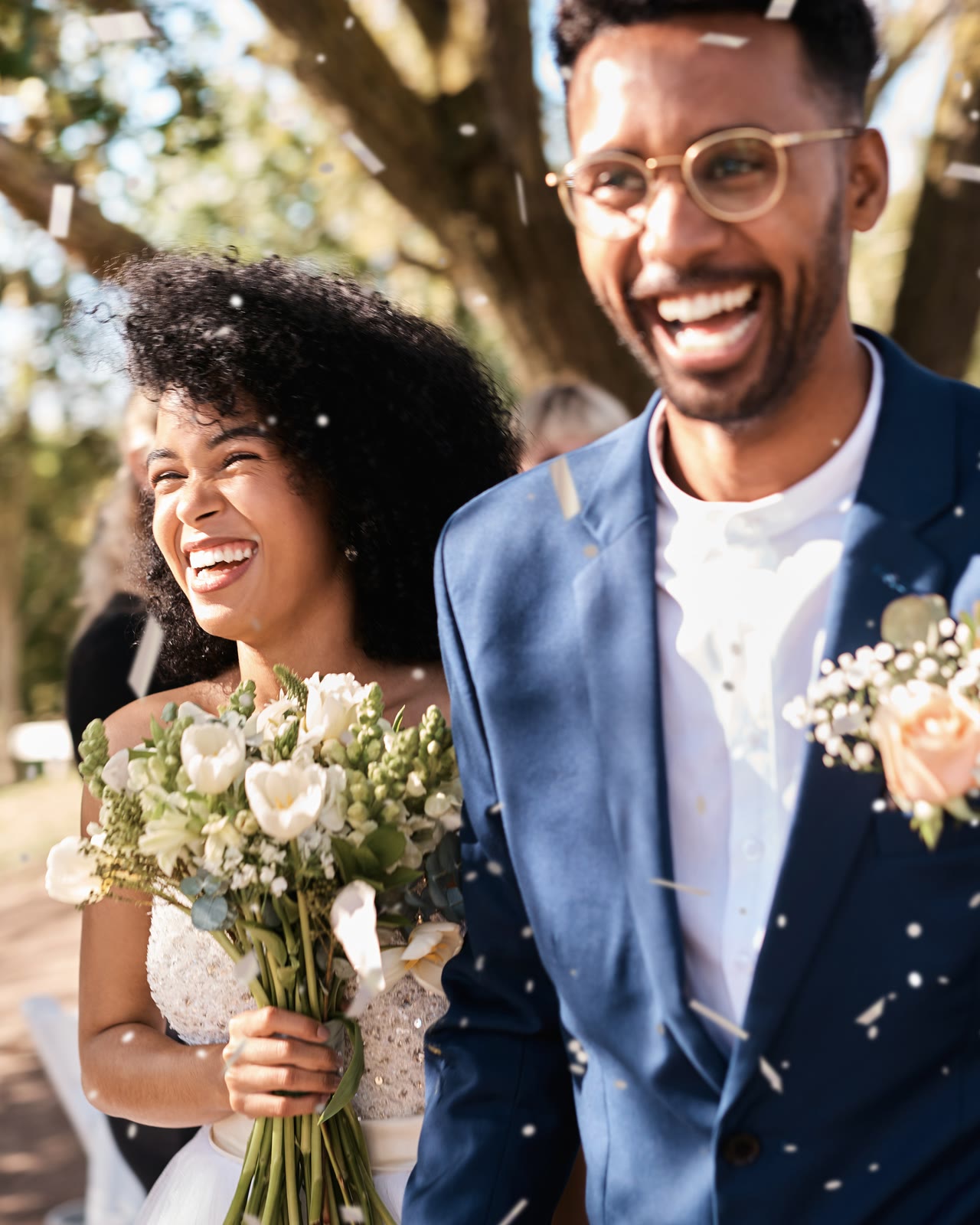 A couple laughing through falling confetti under the trees, a bouquet in her hands.