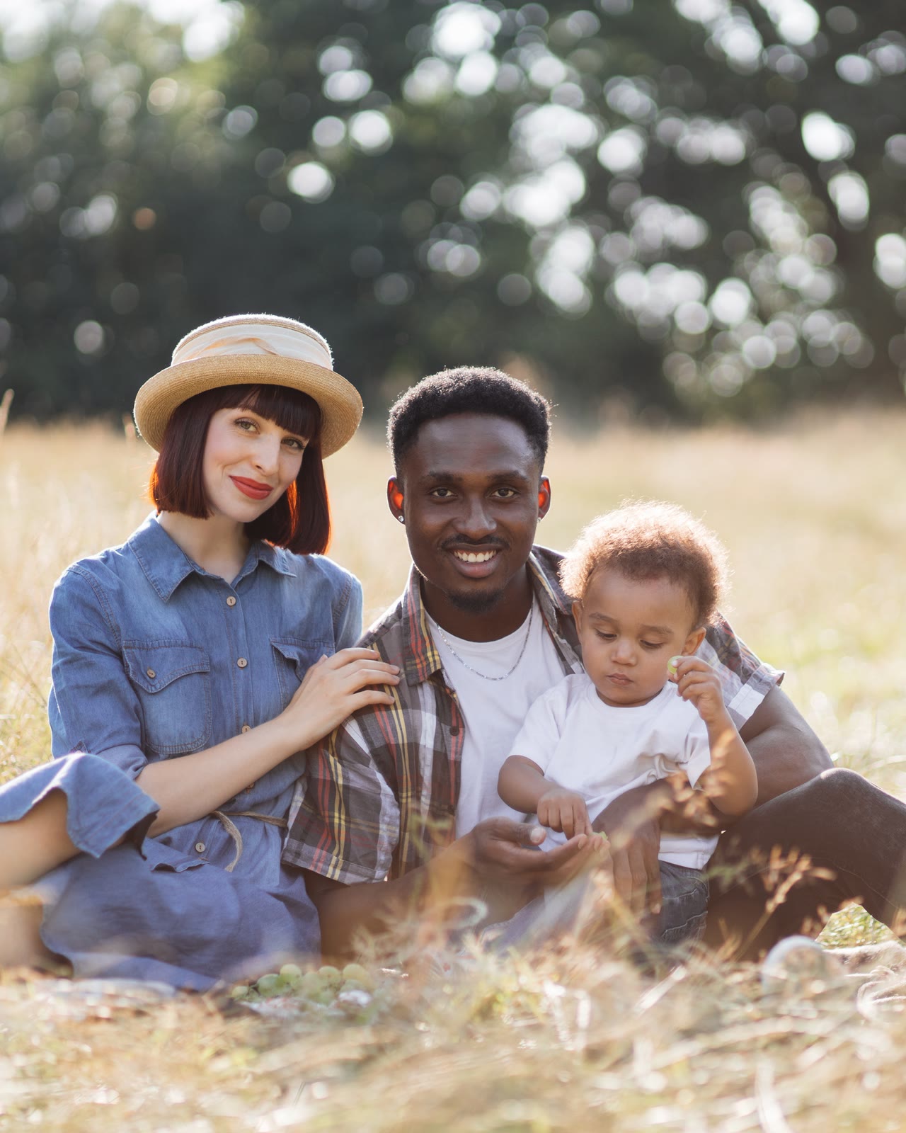 A couple with a toddler sitting in dry golden grass, soft light behind them.