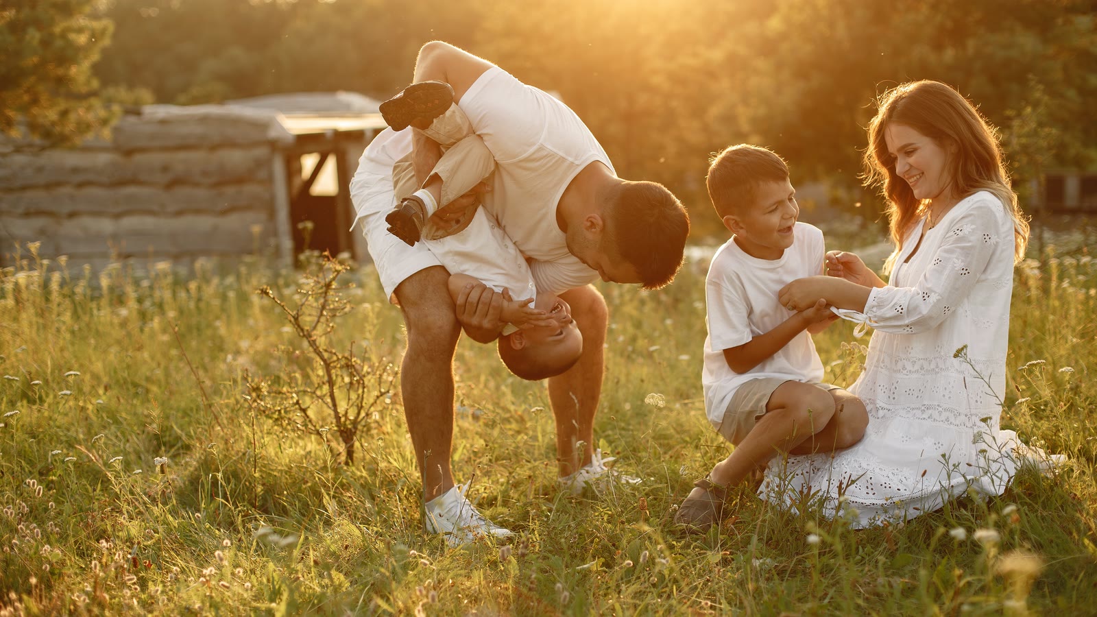 A family playing in tall dry grass at sunset, a child swung upside down, a boy and his mother laughing.