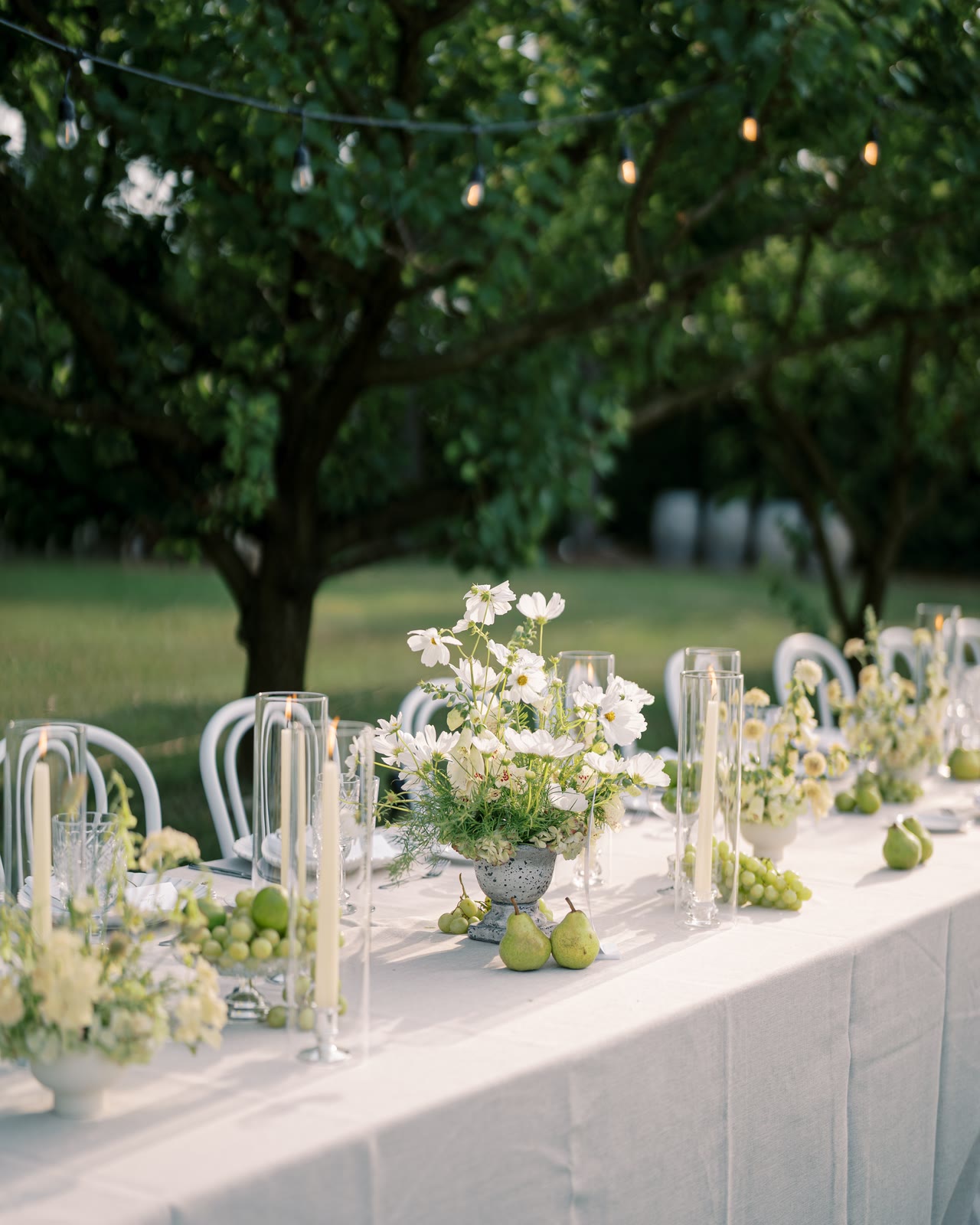 A long white table set under orchard trees with pears, white flowers and taper candles.