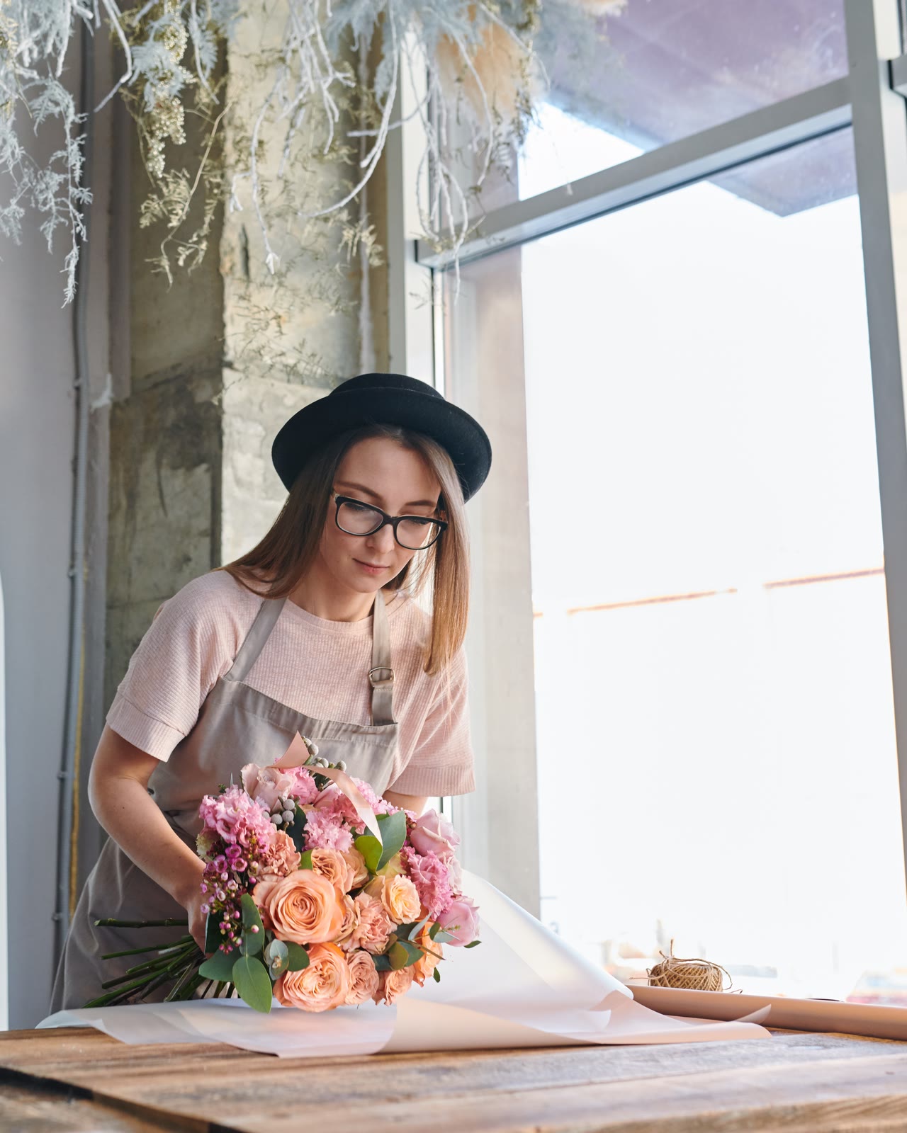 A florist wrapping a bouquet of roses at a table by a tall window.